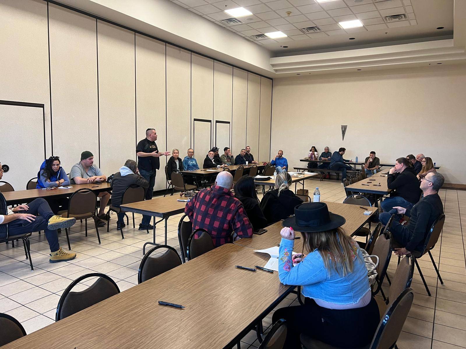 People seated at long tables in a meeting room while one person stands at the front addressing them