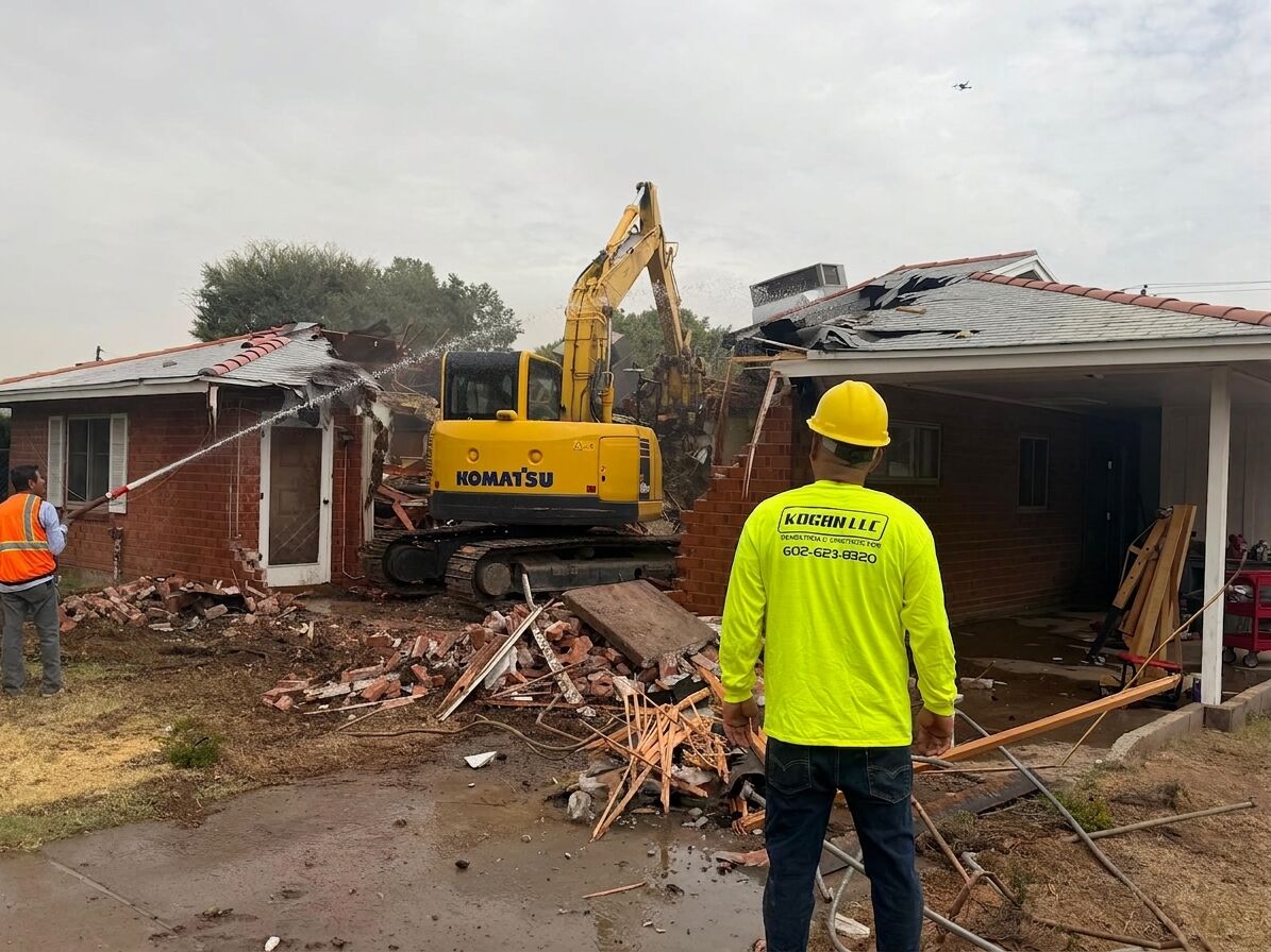 A Kogan LLC crew member in a hard hat and branded high-visibility shirt watching a Komatsu excavator tear into a brick house in Phoenix, Arizona