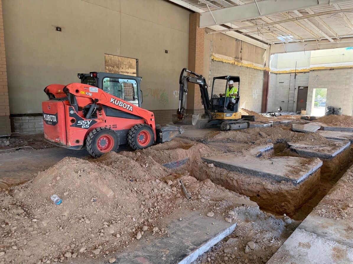 A Kogan LLC operator in a hard hat and high-visibility vest running a mini excavator beside a Kubota skid steer during a commercial interior strip-out in Phoenix, Arizona